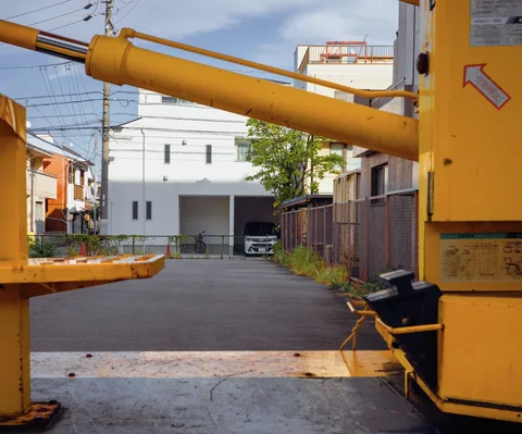 A yellow mechanical arm crosses an open paved entrance between low buildings beneath overhead wires.