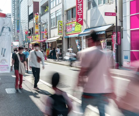 Pedestrians, some blurred by movement, fill a shopping street lined with colorful storefront signs.