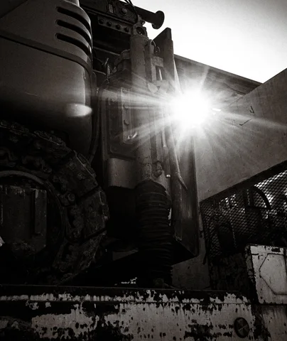 Bright sunlight shines between bulky weathered machine components above a chipped metal platform in black and white.