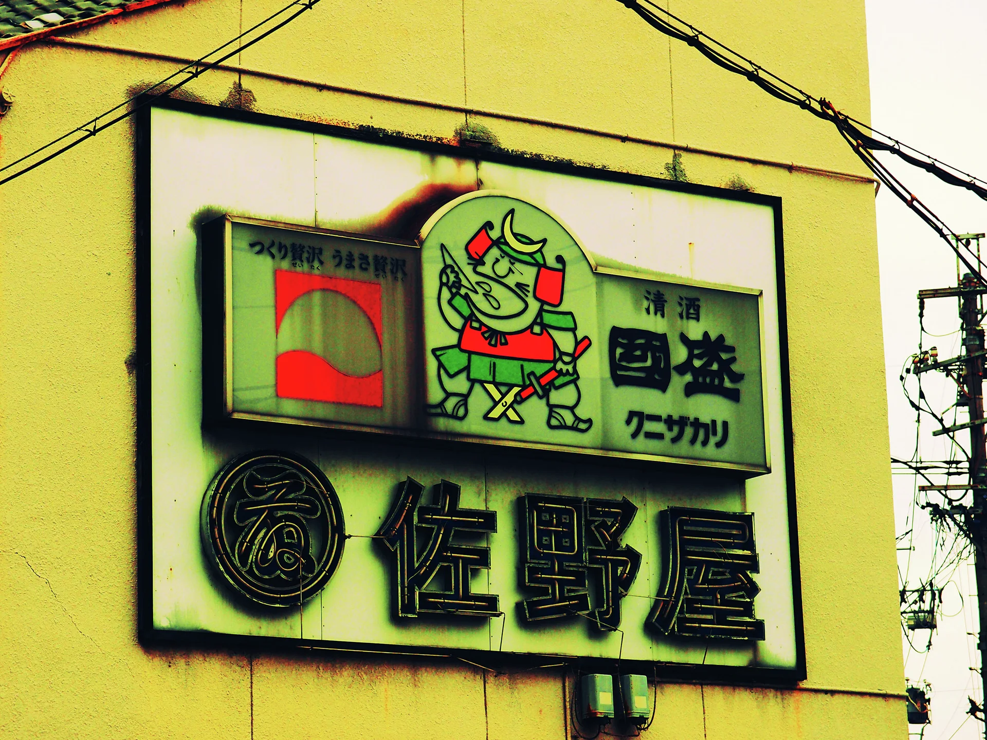 A large rectangular sign combines raised Japanese characters and a cartoon figure against a yellow building.