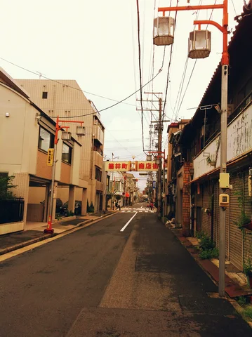 An empty narrow street runs between low buildings beneath overhead wires and paired hanging street lanterns.