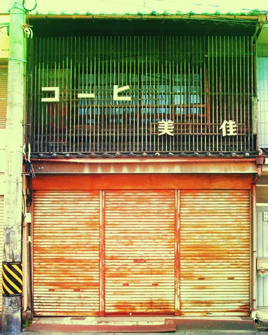 Three rust-streaked shutters close a narrow storefront beneath a barred upper window with pale Japanese lettering.