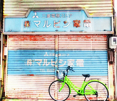 A bright green bicycle leans against a weathered red, white, and blue shutter beneath a pale blue sign.
