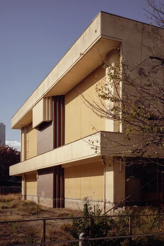 Broad pale concrete ledges project from a tan building beside sparse trees and low fencing.