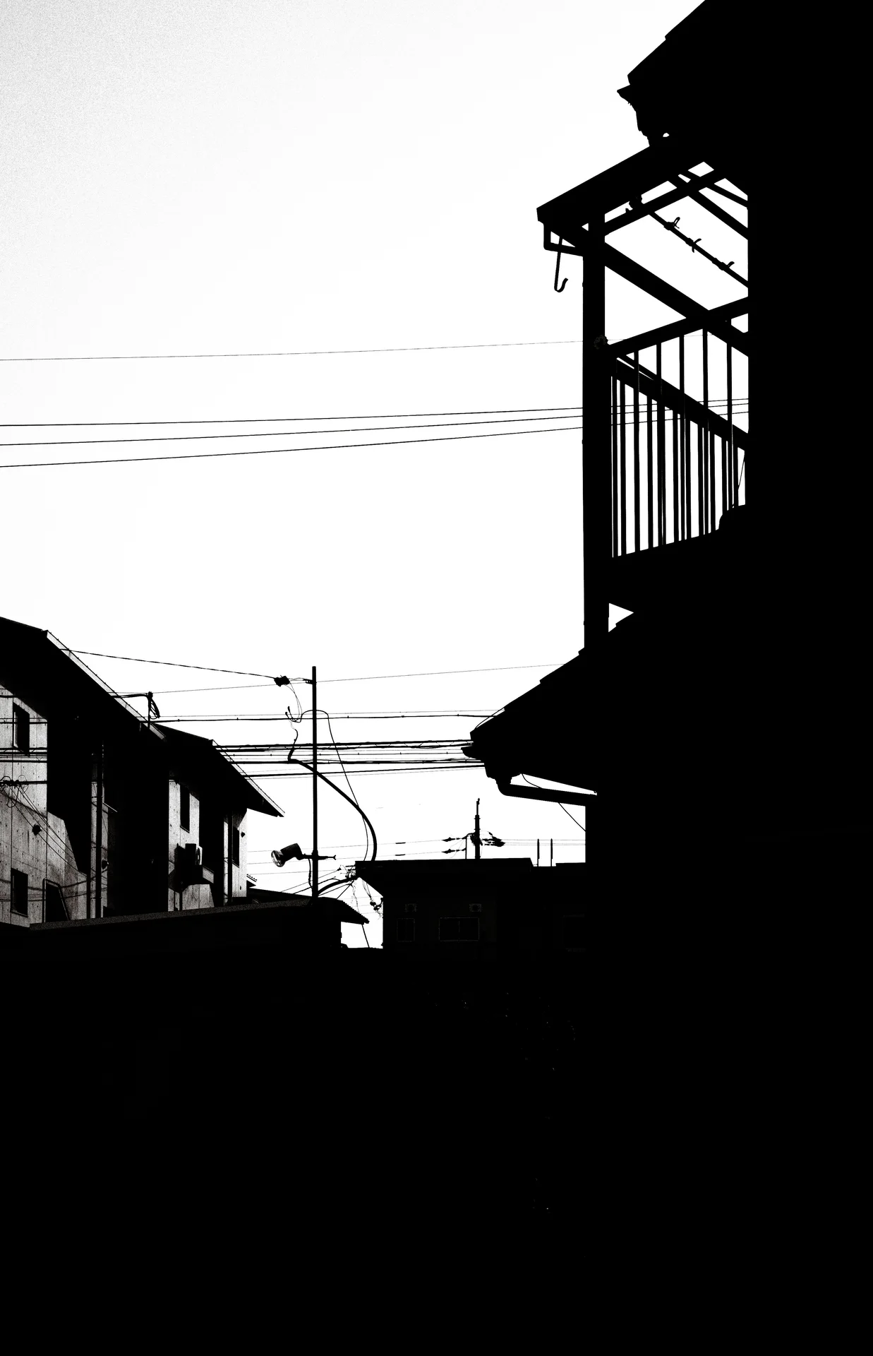 Dark rooftops and a projecting balcony silhouette frame a pale sky crossed by horizontal wires.