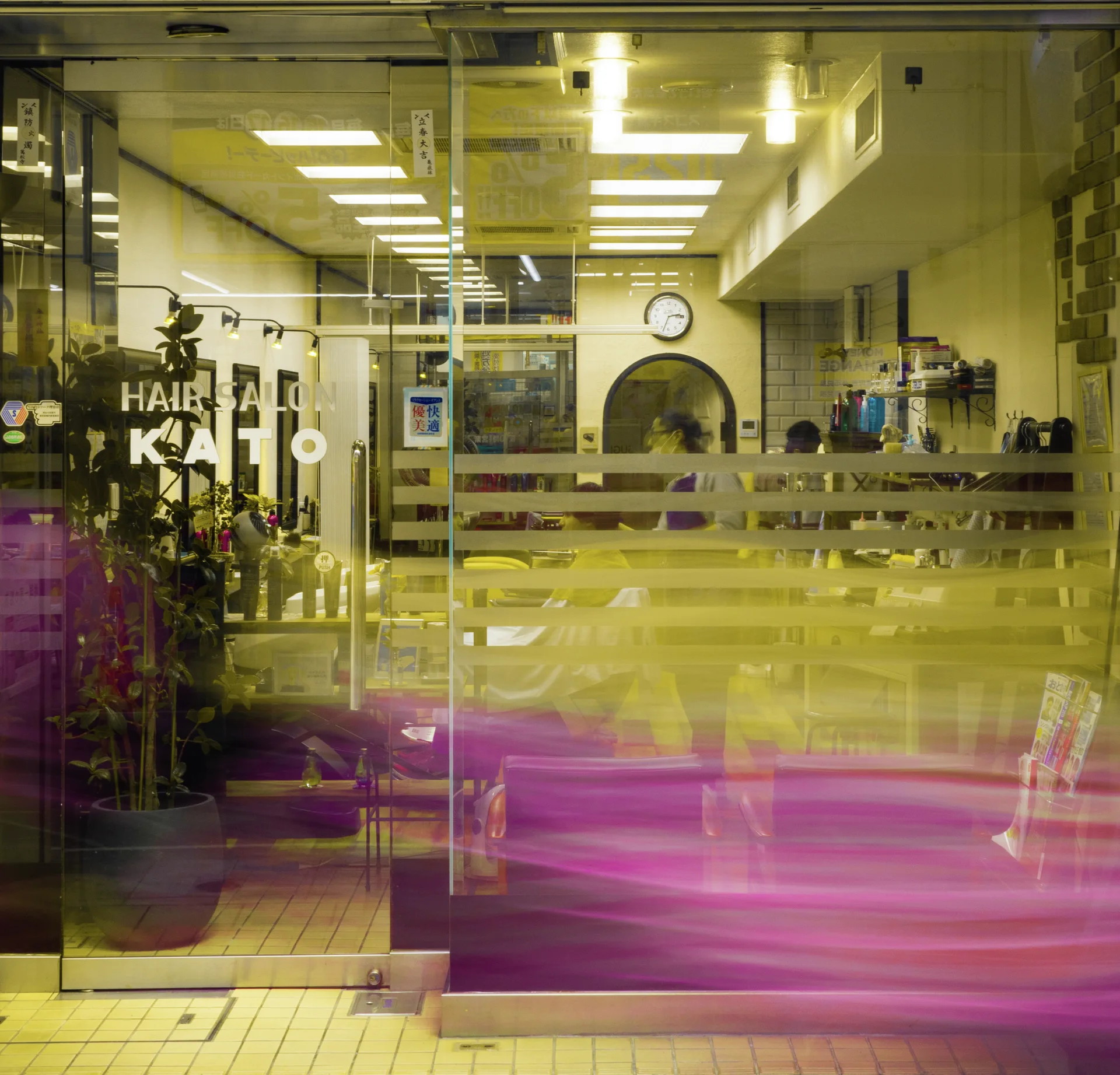 A glass salon entrance reveals rows of ceiling lights while a pink blur crosses its lower panes.