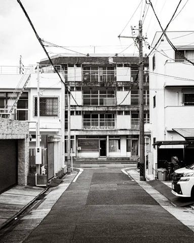 A narrow residential lane ends at an old multi-story facade beneath tangled wires in black and white.