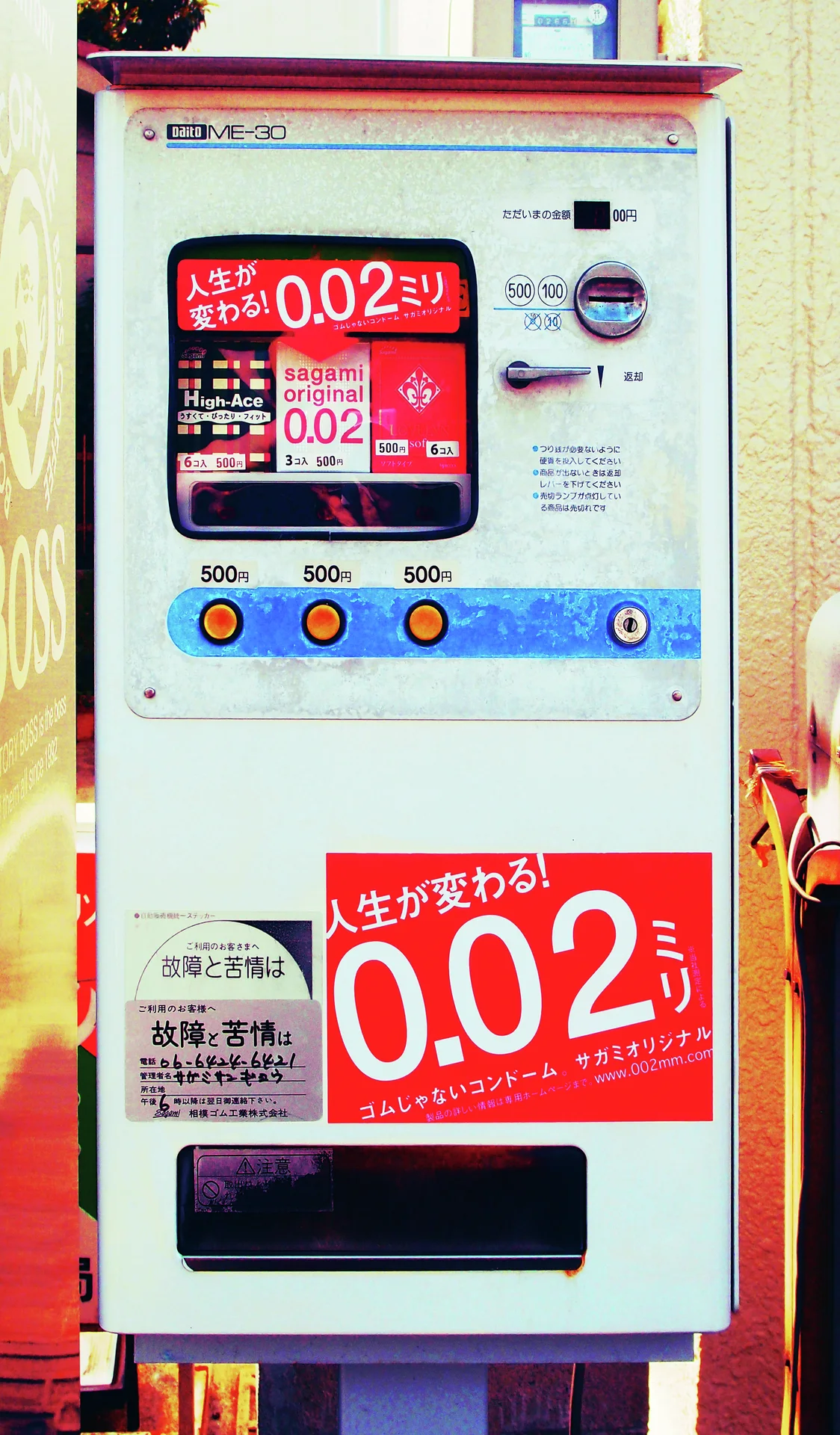 A narrow pale vending machine has red promotional panels, a blue button strip, and a dark lower opening.