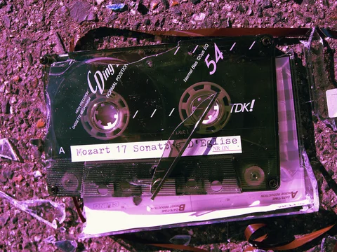A black cassette labeled with handwritten text lies among broken plastic pieces on pink-tinted pavement.