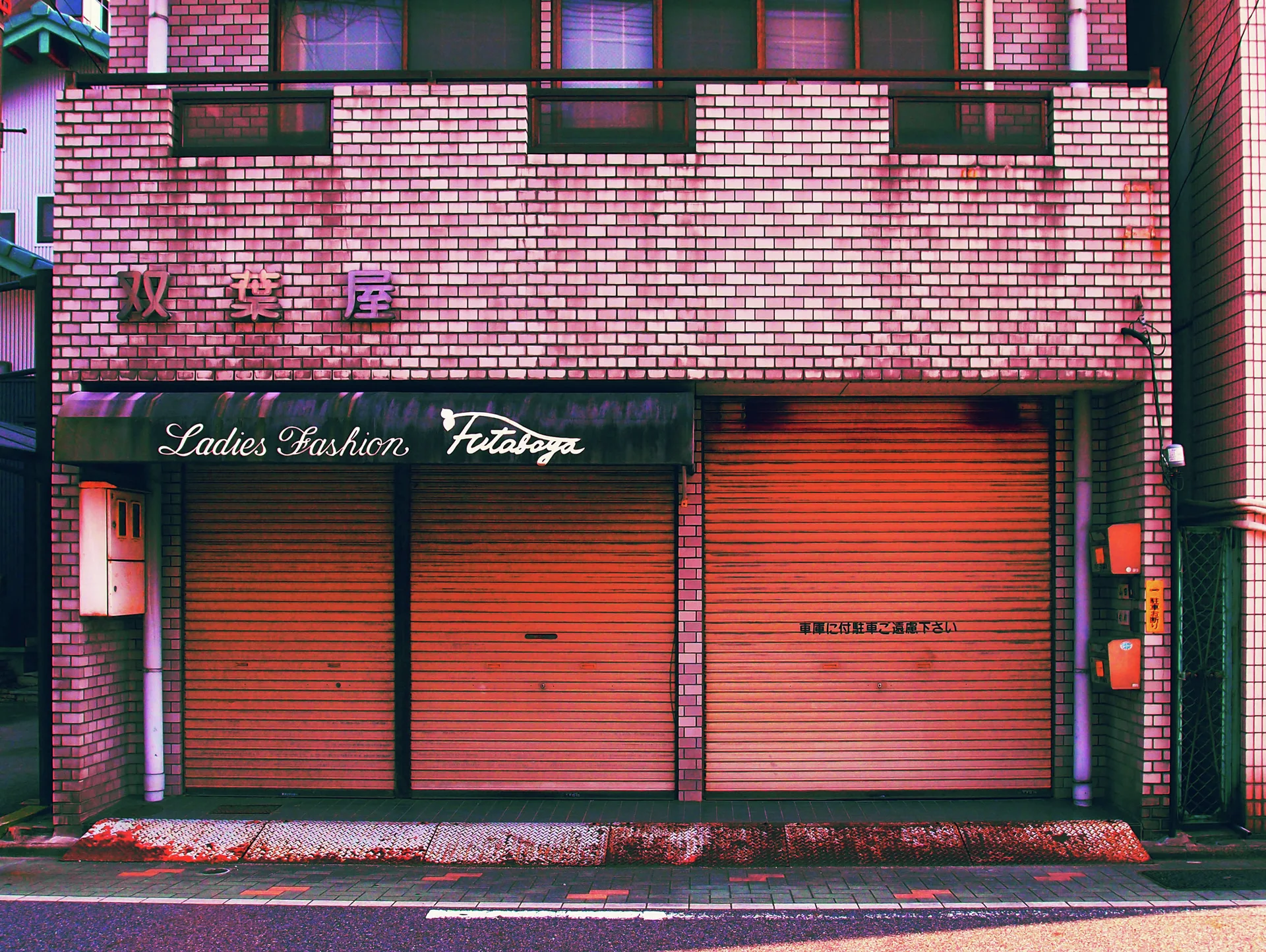 Closed pink shutters line a tiled clothing shop beneath a dark awning with cursive white lettering.