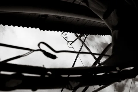 Dark cables and a television antenna cross bright sky beneath a corrugated roof in black and white.