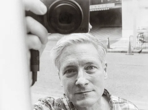 A man's face and a camera held above him appear together in a black and white reflection.