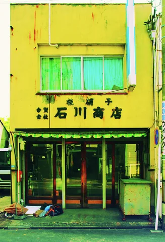 A bright yellow narrow shop has green upper windows and Japanese lettering above its glass entrance.