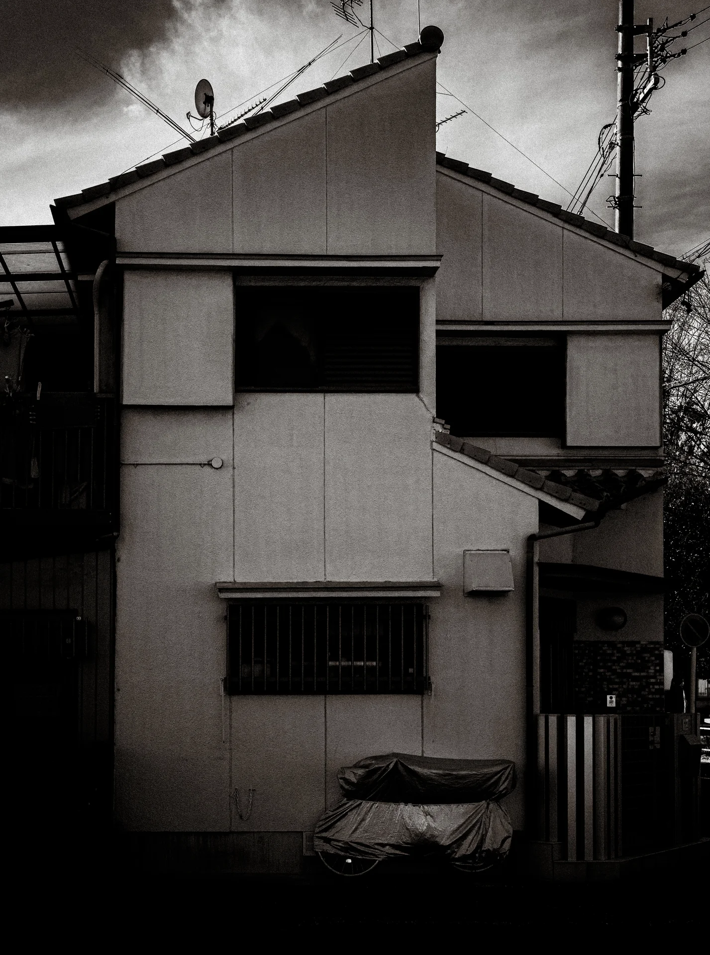 A narrow angular house with dark windows stands beneath cloudy sky in black and white.