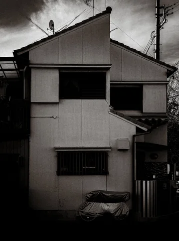 A narrow angular house with dark windows stands beneath cloudy sky in black and white.