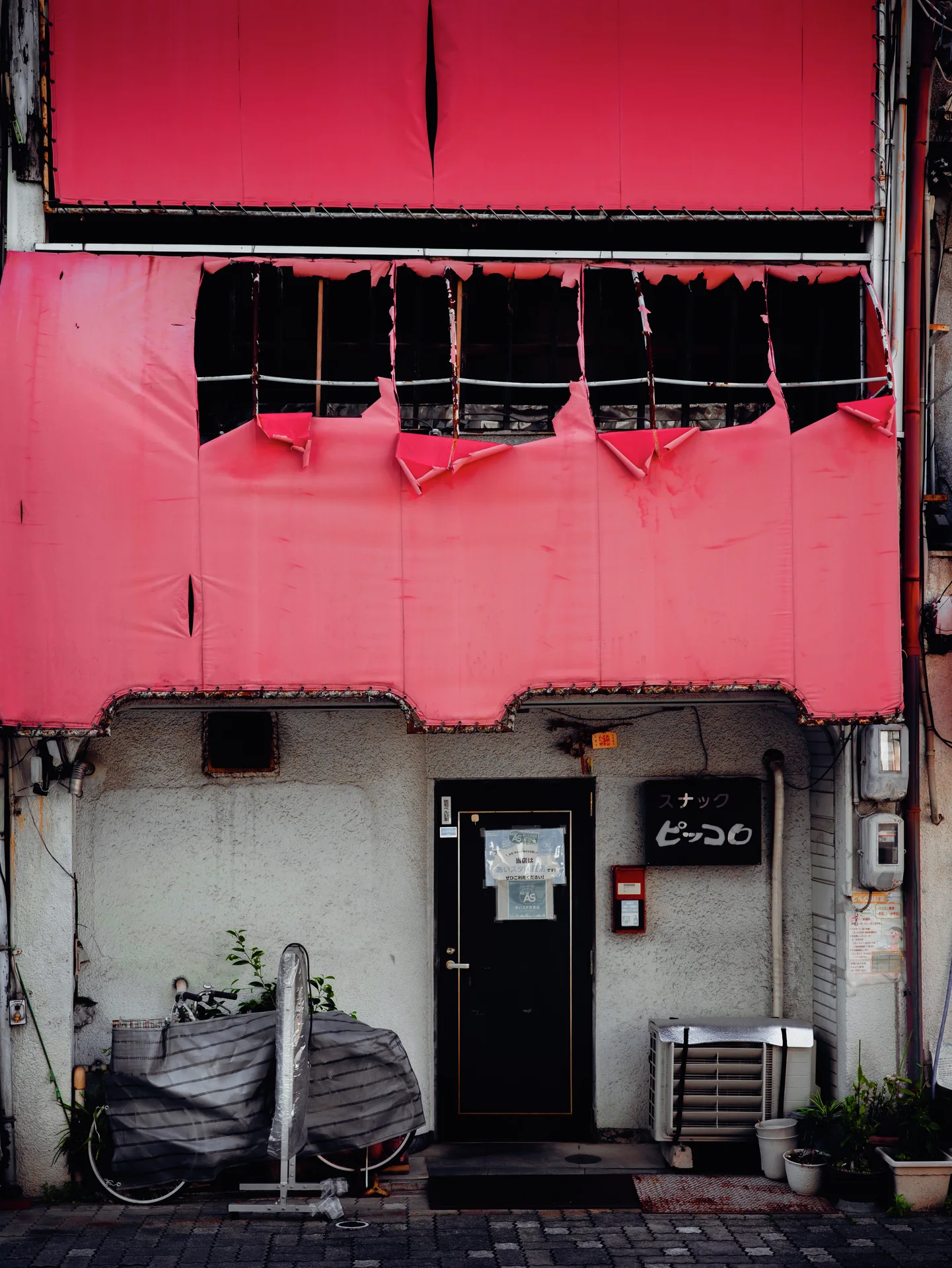 A damaged pink upper facade rises above a dark entrance and covered bicycle against a gray lower wall.