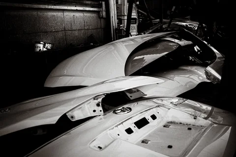 Detached pale vehicle panels lie stacked in a dark workshop in a black and white view.