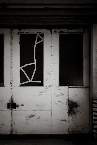 A cracked window sits in a peeling white door beside a dark opening in monochrome.