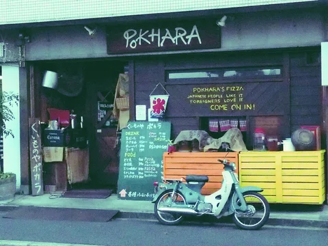 A pale turquoise scooter parks outside a dark restaurant with chalkboard menus and yellow wooden barriers.