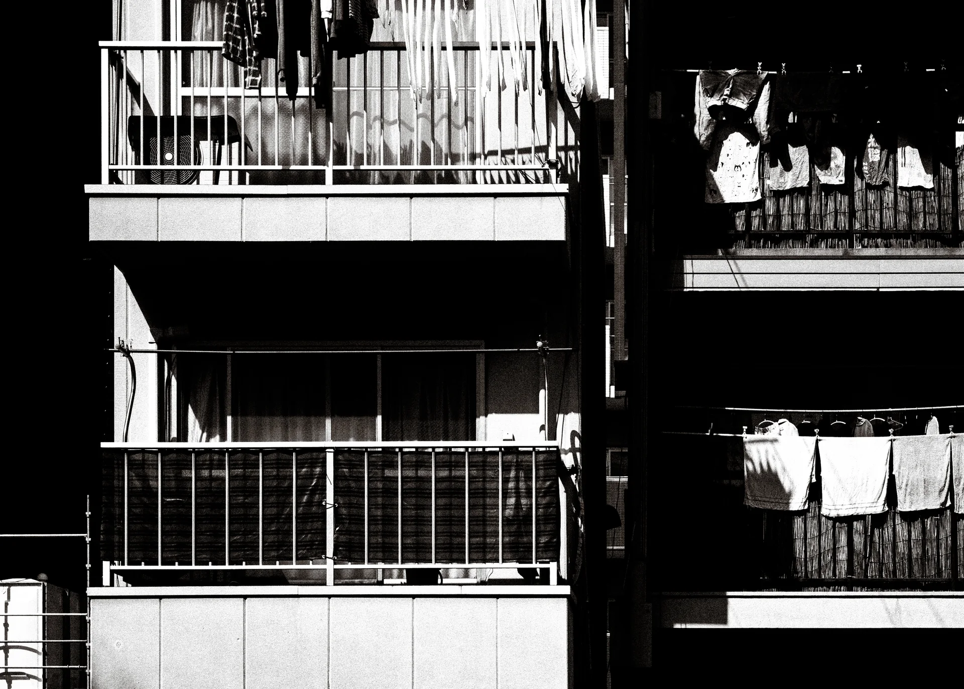 Sunlit laundry hangs from neighboring apartment balconies separated by deep shadows in a black and white view.