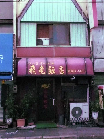 Gold Japanese lettering spans a curved maroon awning above a narrow restaurant entrance and potted plants.