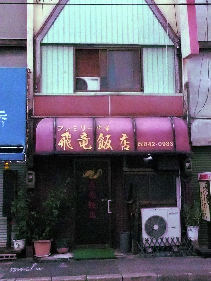 Gold Japanese lettering spans a curved maroon awning above a narrow restaurant entrance and potted plants.