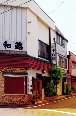 A narrow corner restaurant has pale upper siding, a red awning and stone-patterned lower walls.