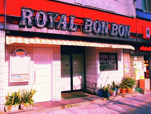 Large Royal Bonbon lettering crowns a pale pink storefront with a striped awning and potted plants.