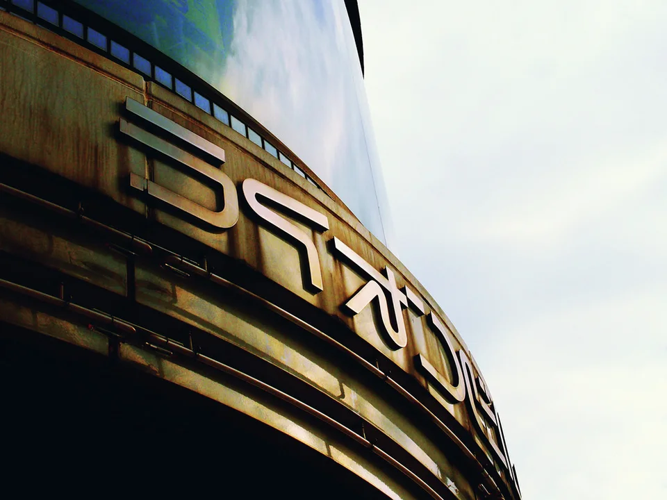 Large pale stylized lettering curves across a dark tiled facade viewed steeply upward against a bright sky.