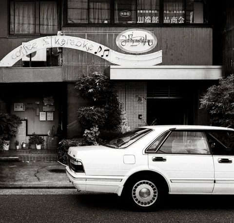 A white sedan is parked beneath curved storefront signs and upper windows in black and white.