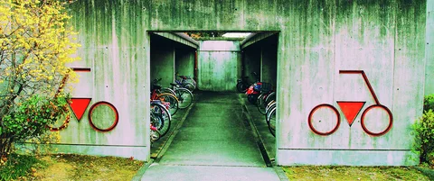 Red bicycle symbols flank an open passage lined with parked bicycles in a green-stained concrete wall.