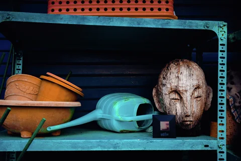A weathered carved face shares a turquoise metal shelf with plant pots and a green watering can.