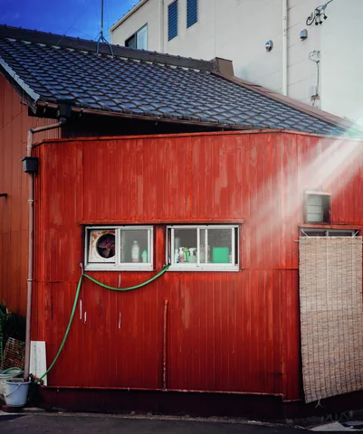 Sunlight flares across a red corrugated wall with two small windows and a hanging green hose.