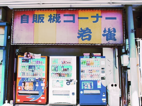 Three colorful drink vending machines stand beneath a faded pink and yellow Japanese sign beside a small white bin.