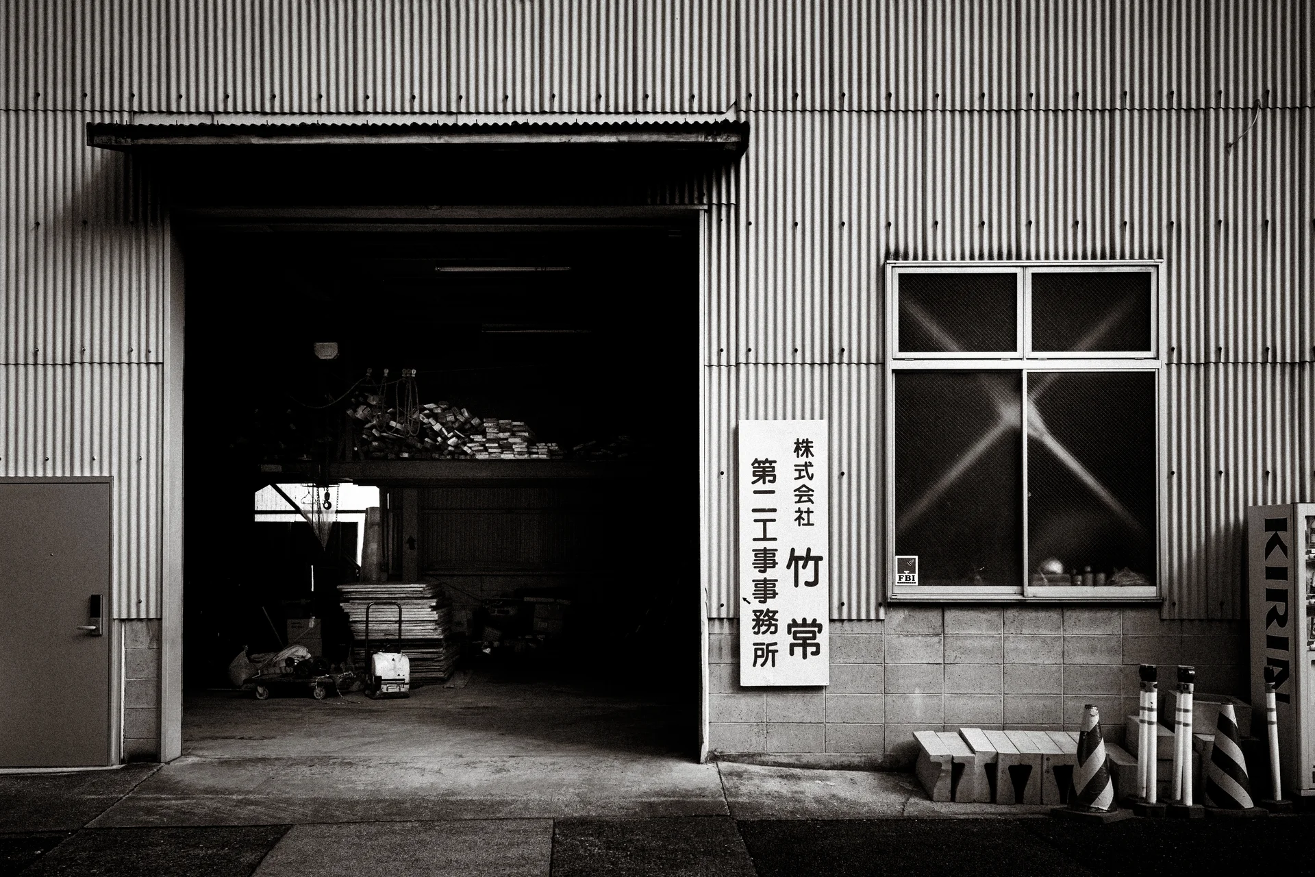 A broad open workshop doorway reveals stacked materials beside a cross-braced window in black and white.