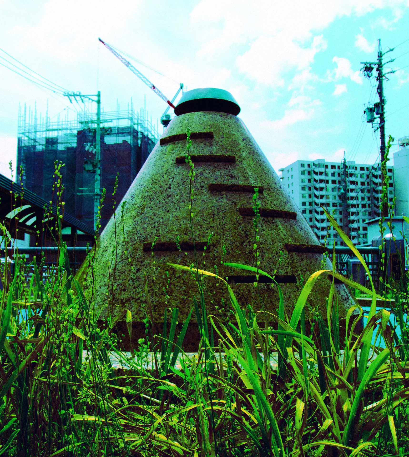 A rough cone-shaped structure with projecting metal steps stands among tall grass before distant apartment buildings.