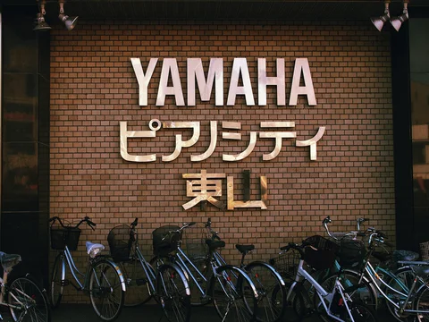 Many bicycles park beneath large pale YAMAHA letters and Japanese signage on a brick-patterned wall.