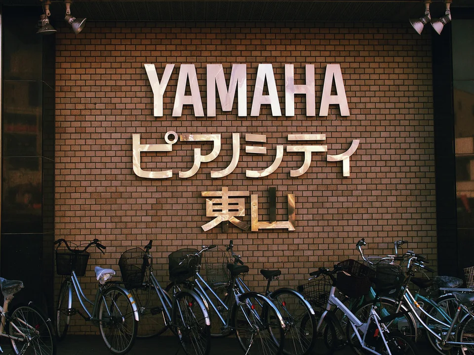 Many bicycles park beneath large pale YAMAHA letters and Japanese signage on a brick-patterned wall.