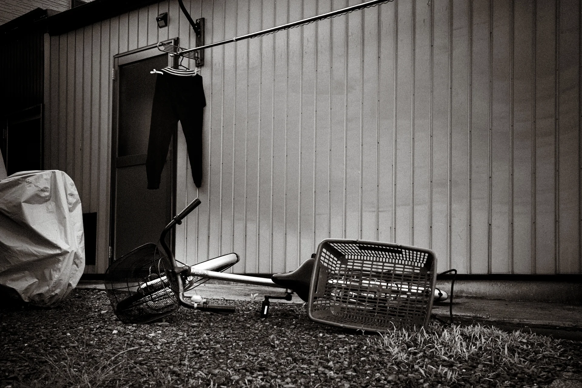A fallen bicycle lies beneath trousers hanging beside corrugated siding in a black and white view.