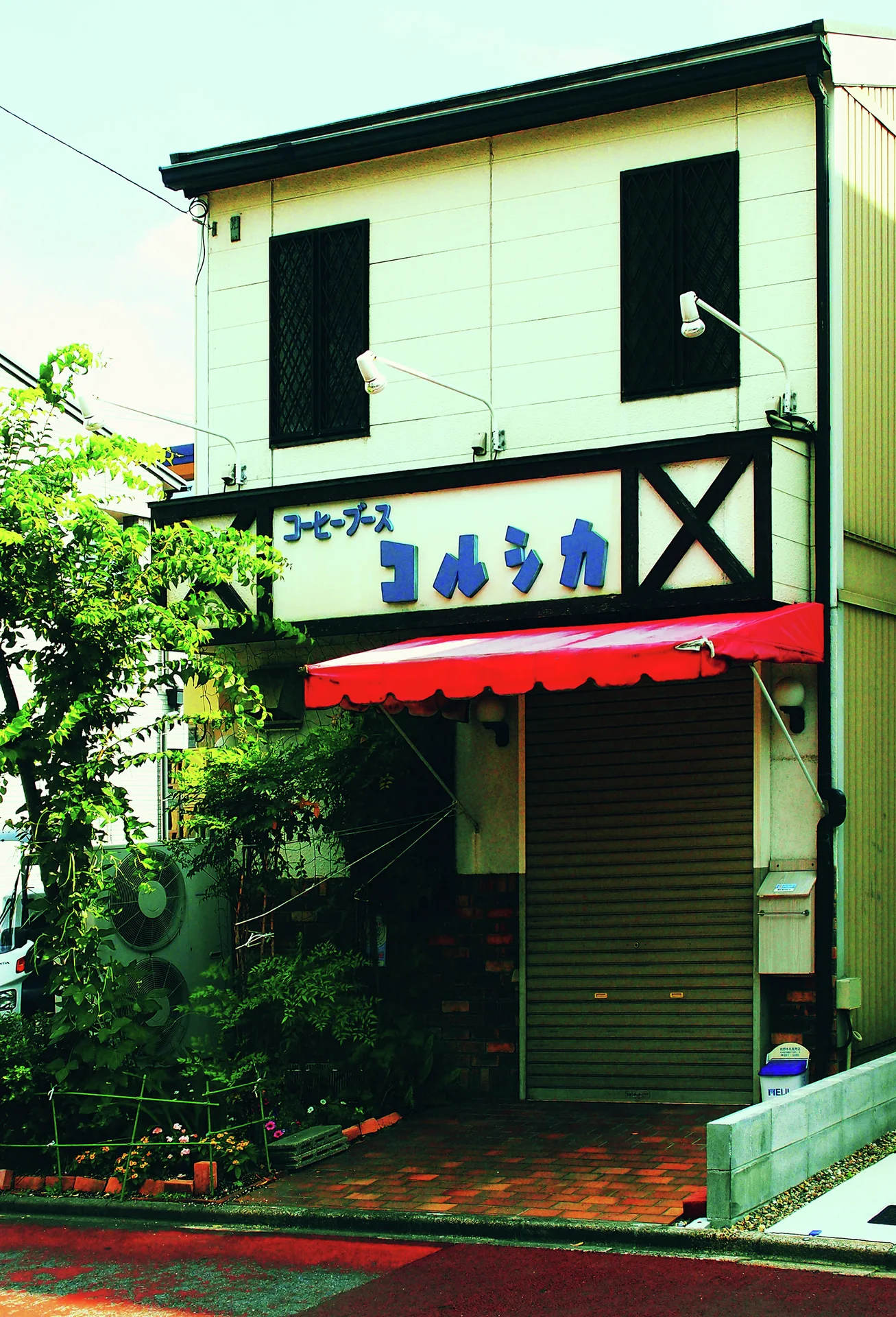 A red awning shades a narrow closed shop with dark upper windows and surrounding greenery.
