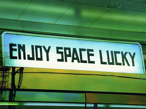 Angular black ENJOY SPACE LUCKY lettering spans a glowing pale sign beneath a green-toned ceiling.