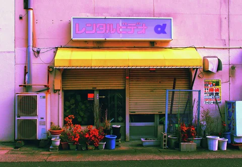 A yellow awning shades a pink storefront with partly lowered shutters and pots of red flowers.