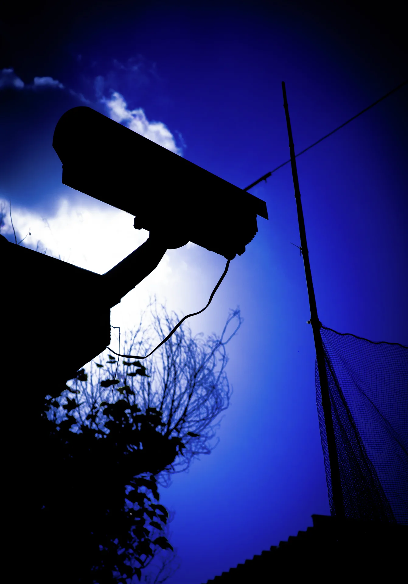A dark projecting fixture silhouettes against white clouds and deep blue sky beside a slender antenna.