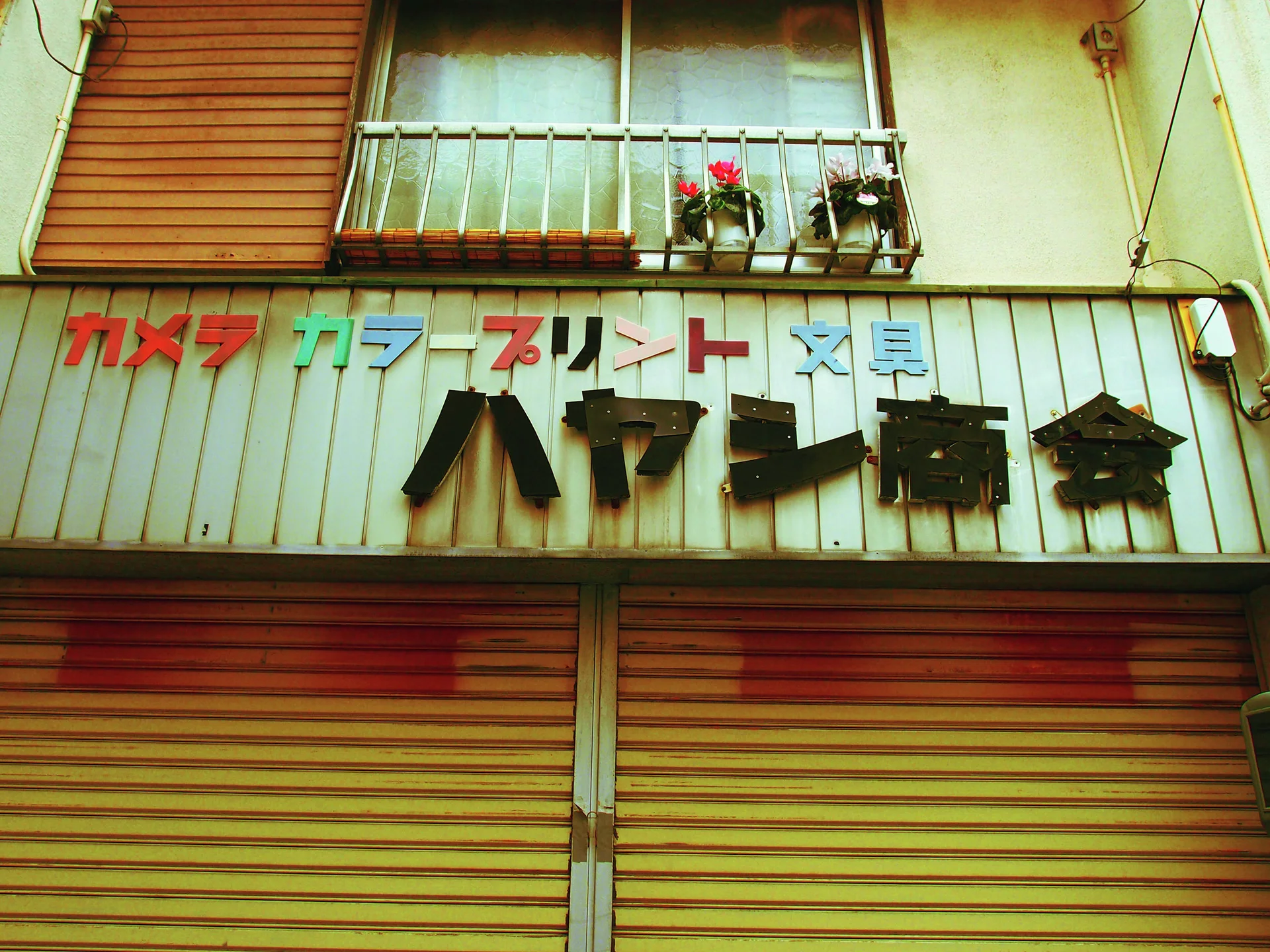 Multicolored camera-shop lettering hangs above striped shutters beneath a balcony with small flowerpots.