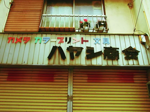 Multicolored camera-shop lettering hangs above striped shutters beneath a balcony with small flowerpots.
