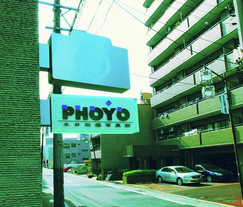 A projecting turquoise sign overlooks a quiet street beside long apartment balconies and parked cars.
