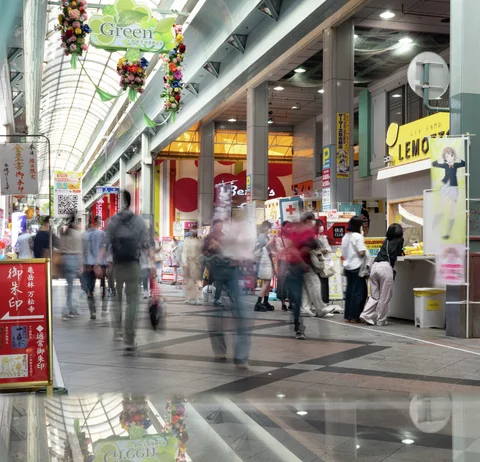 Blurred pedestrians move through a covered shopping arcade beneath hanging decorations and brightly colored shop signs.