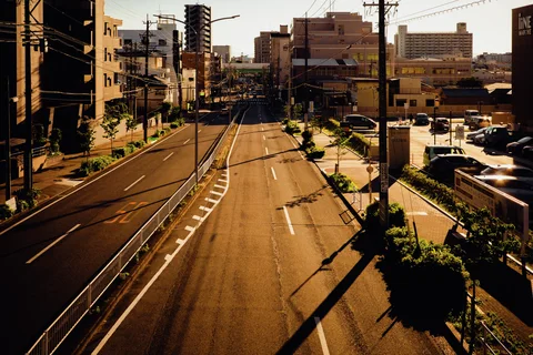 A broad divided road stretches between city buildings under low sunlight with long utility-pole shadows.