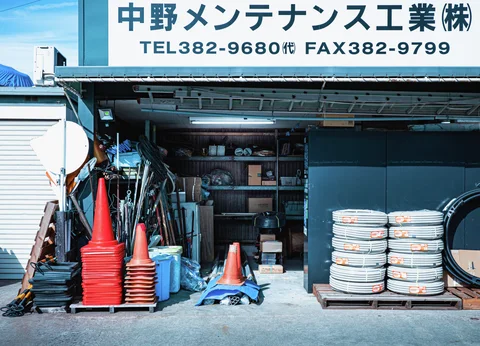 Stacks of orange traffic cones and coiled white tubing flank an open workshop filled with supplies.