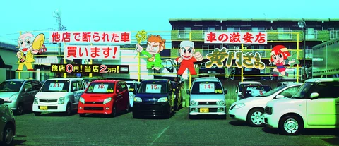 Rows of compact cars display windshield prices beneath large colorful signs with cartoon figures.